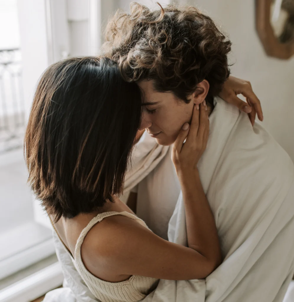 A young couple in a tender embrace in the bedroom. The woman has short, dark hair; the man has curly hair and is wrapped in a white blanket. They gently touch each other's faces.
