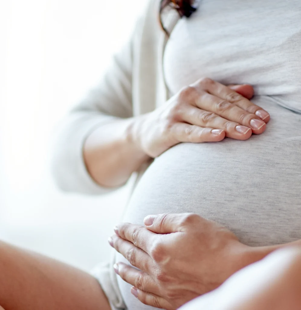A close-up of a pregnant woman in light-colored clothing gently placing both hands on her round baby bump.