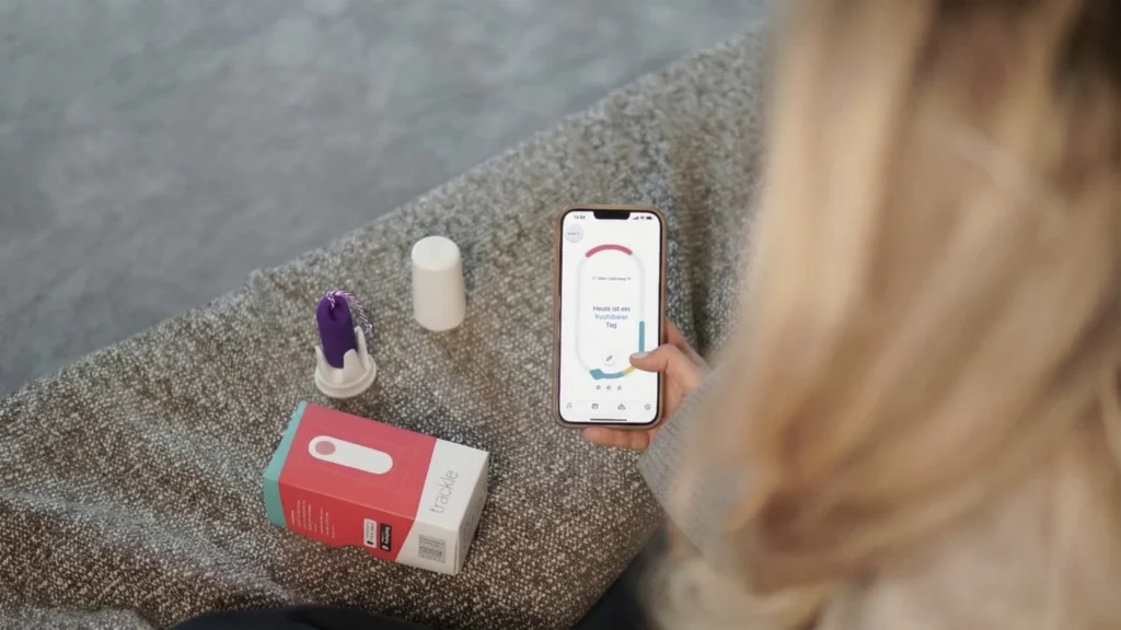 A view over the shoulder of a woman holding her smartphone. The trackle app’s display shows the message “Today is a fertile day.” Next to her, on a textured surface, lie the purple trackle sensor in its holder and the product packaging. The image illustrates how digital cycle tracking works in everyday life.