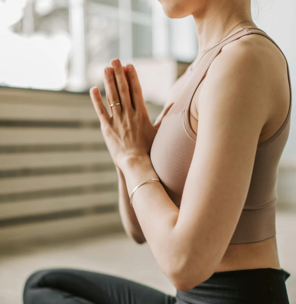 A side view of a woman in a yoga pose, with her hands pressed together in front of her chest in a greeting (Namaste). She is wearing a beige sports top.